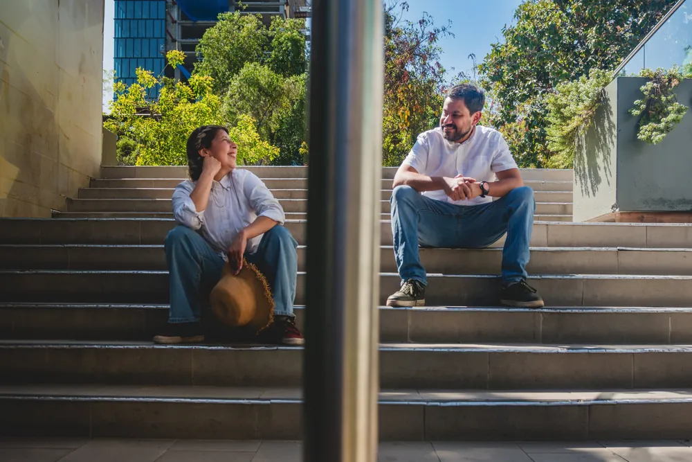 Sayda y Marco sentados en una escalera, separados por un poste, se miran y se ríen