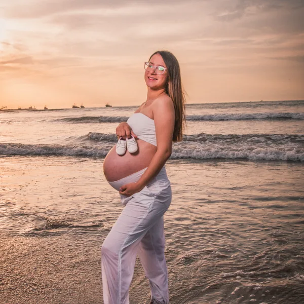 Mujer embarazada en la playa al atardecer, con unos zapatitos de bebé en la mano