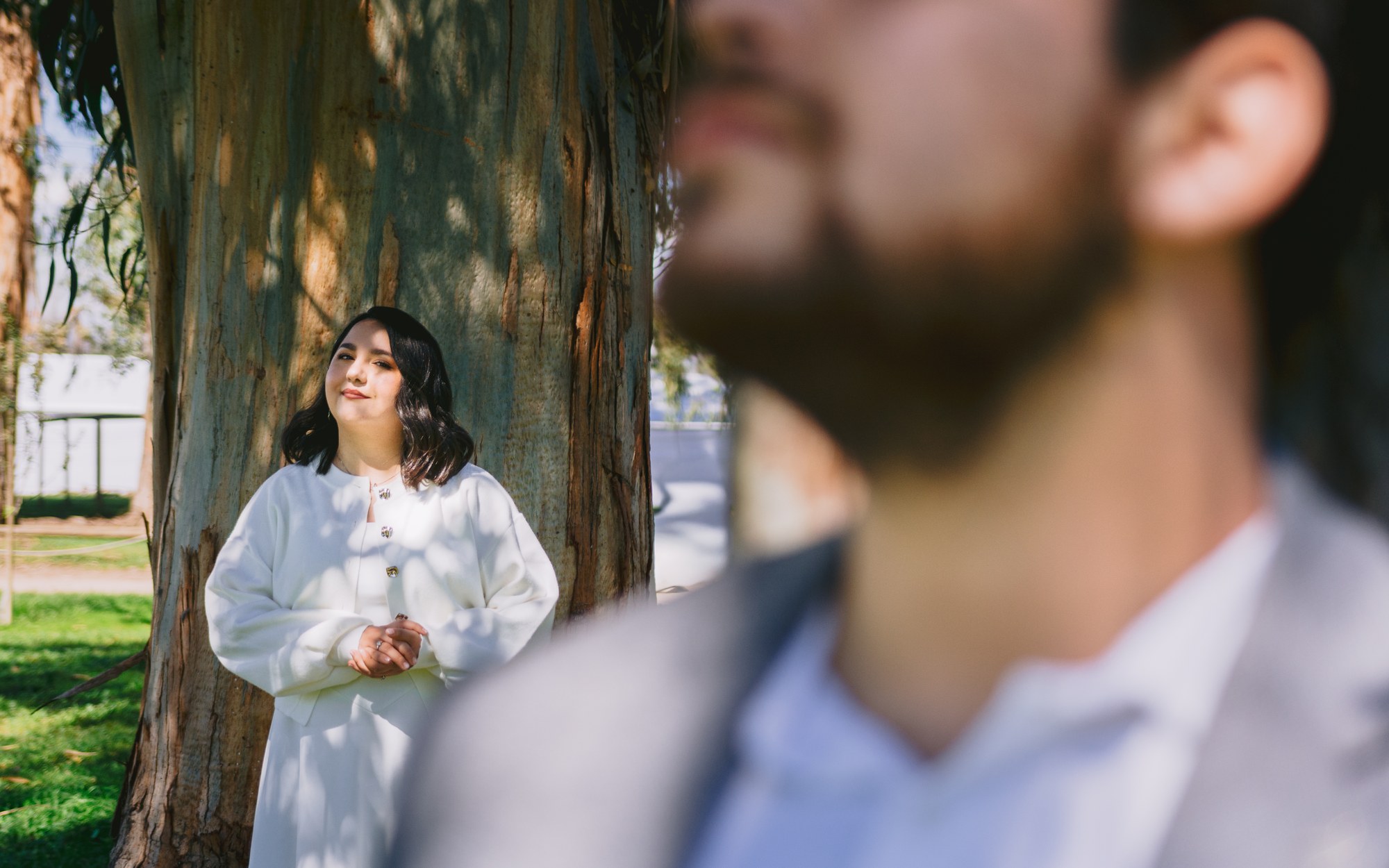 Novia apoyada en un árbol, con el novio desenfocado en primer plano
